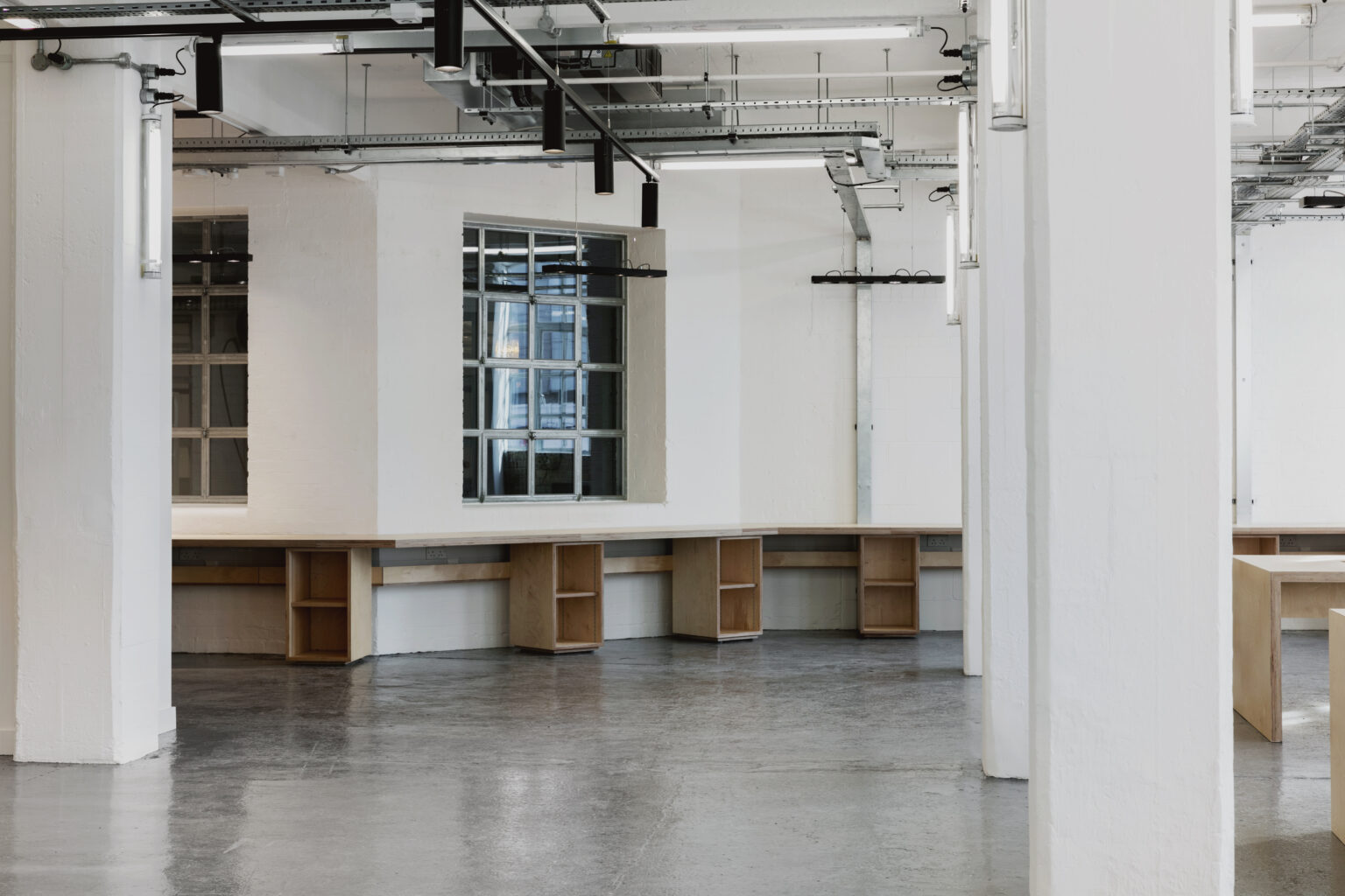 Plywood workbenches beneath steel-framed windows and exposed brick at the Tea Building